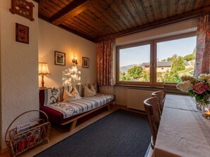 A cozy room with a wooden ceiling, a window with a view of greenery, a sofa with cushions, a table with chairs, and a radiator. - Spacious apartment with beautiful mountain views (Zell am See)