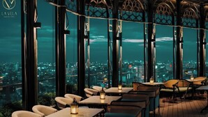 Een eetruimte met tafels en stoelen, grote ramen met uitzicht op de stad, en een decoratief plafond.
