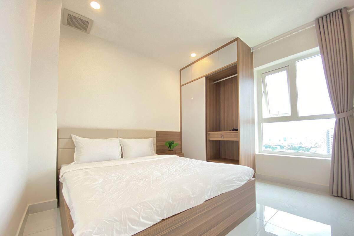 Une chambre d’hôtel moderne avec un grand lit, une armoire intégrée, une petite plante et une vue sur la ville par la fenêtre.