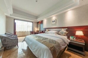 A hotel room with a large bed, a desk, a chair, and a nightstand with a lamp.