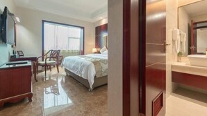 A hotel room with a bed, a desk, a chair, a large window, and a bathroom with a mirror and sink. - Yilong Hotel (Jingdezhen)