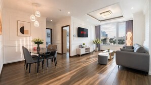 A modern living room with a dining area, a sofa, a TV, and a city view. - Salute de Léman - Luxury Apartment District 3 (Ho Chi Minh City)