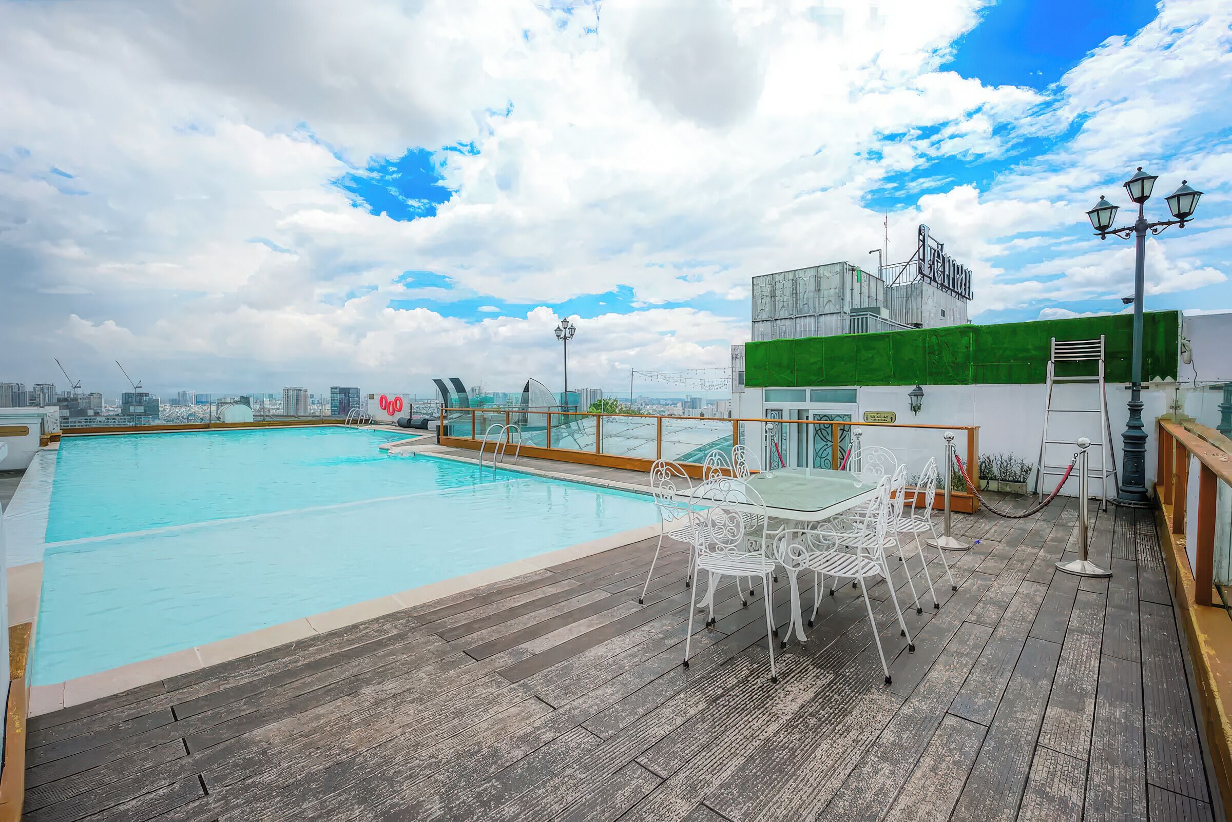 A rooftop pool with a wooden deck, white metal chairs, and a view of the city skyline.