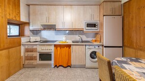 Een kleine keuken met houten kastjes, een magnetron, een koelkast, een wasmachine en een eethoek met rieten stoelen.