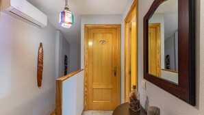 Een gang met een houten deur, een spiegel, een klein tafeltje met decoratie en een luchtverwarmer aan de muur.