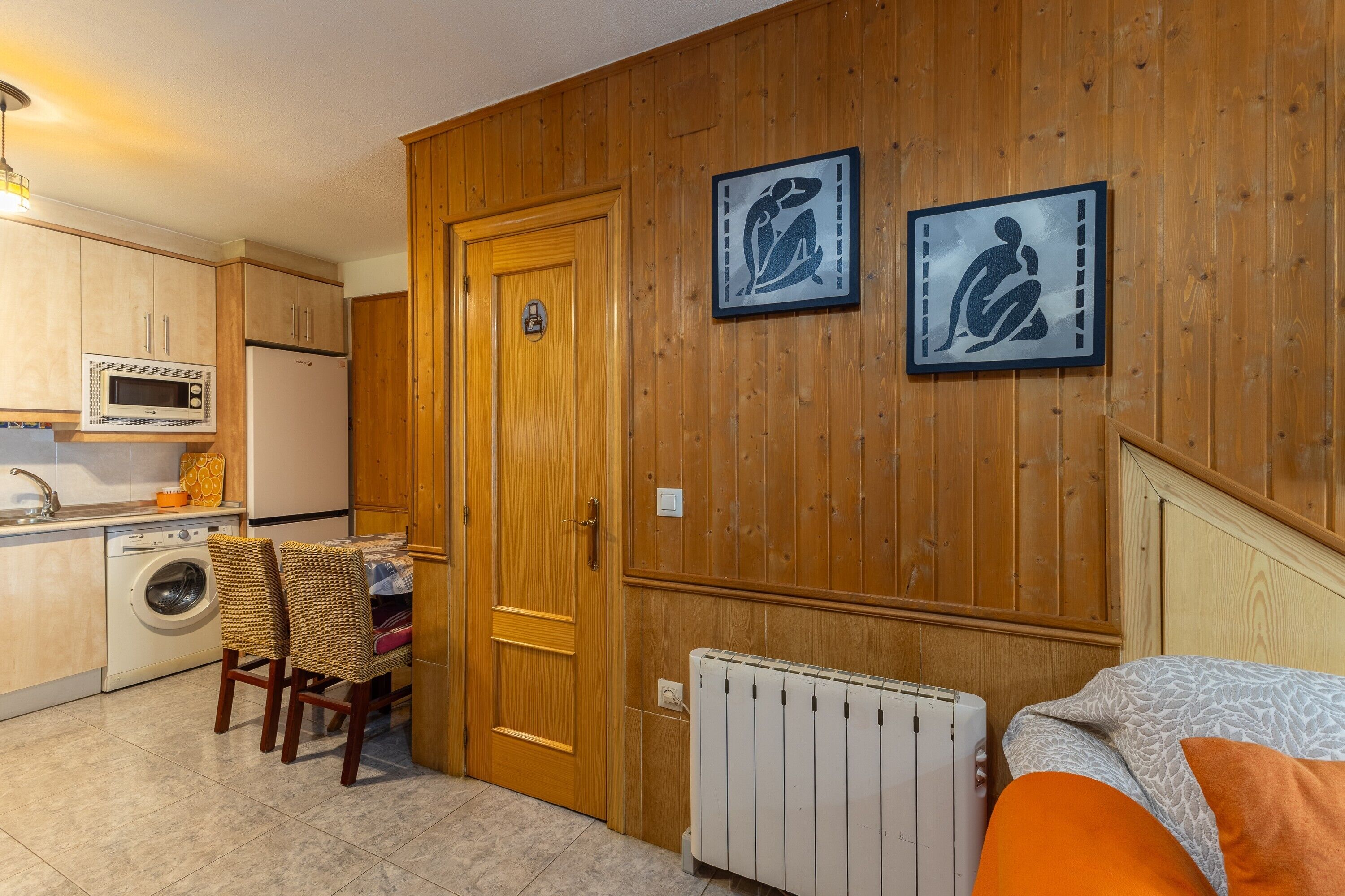Cocina compacta con microondas, lavadora y armarios de madera. Hay dos cuadros enmarcados en la pared y un radiador debajo.