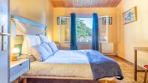 A bedroom with a bed, bedside table, lamp, window with curtains, and a view of greenery outside.