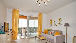 A living room with a sofa, coffee table, TV, and a balcony with yellow curtains. - Apartment 'New Casa Tiberio' with Shared Pool, Wi-Fi and Air Conditioning (Lazise)