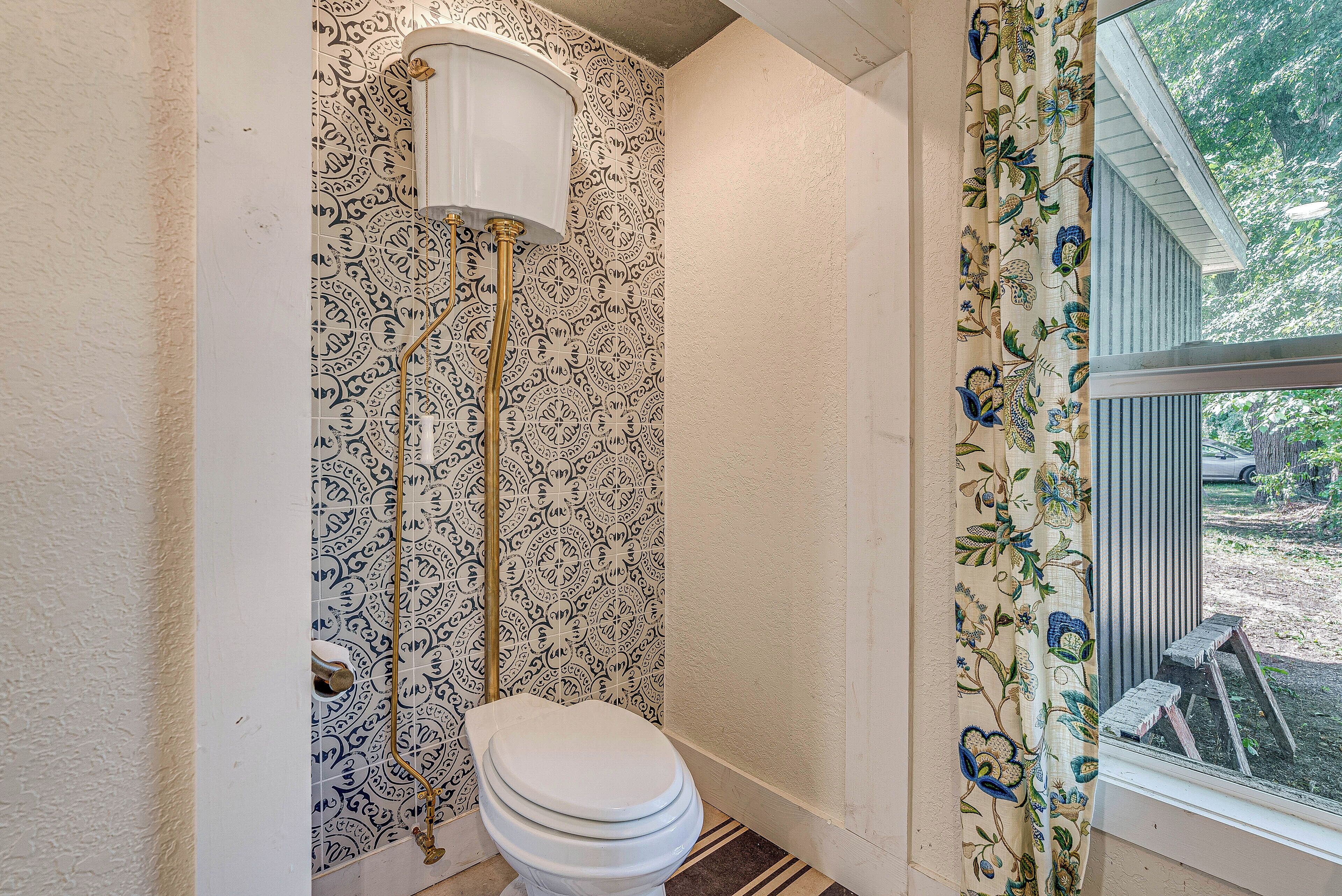 A bathroom with a toilet, a wall-mounted tank, and a window with a view of the outdoors.