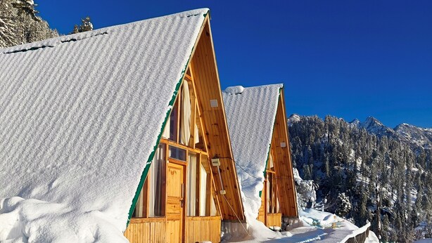 Zwei Holz-Chalets mit verschneiten Dächern, vor einer Kulisse aus schneebedeckten Bergen und wolkenlosem, blauem Himmel.