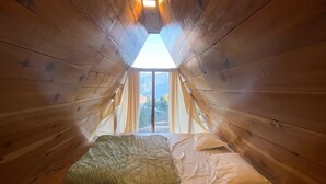 Ein gemütliches Schlafzimmer im Dachgeschoss mit schrägen Holzwänden, einem Bett mit weißer Bettwäsche und Blick auf Bäume durch ein Fenster.