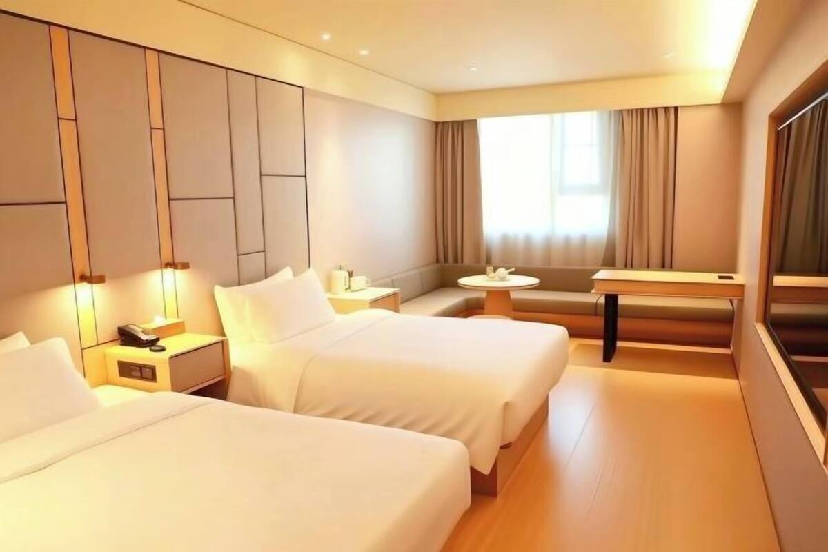 A hotel room with two beds, a small table, a bench, and a large window.