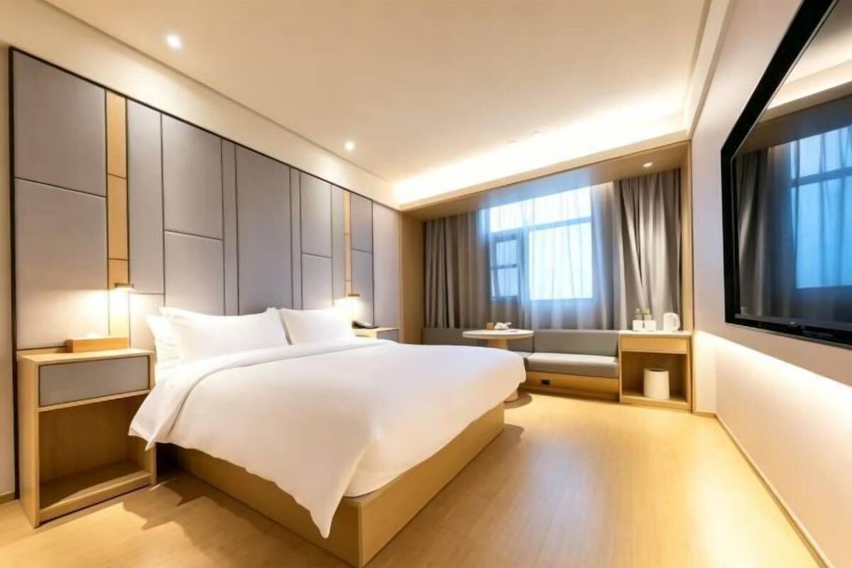 A modern hotel room with a large bed, bedside tables, a window with curtains, and a view of the outside.