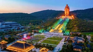 A large statue on a hill with a descending staircase, surrounded by a park and residential buildings.