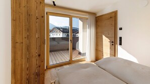 Une chambre avec une armoire en bois, un lit, une porte-fenêtre coulissante donnant sur un balcon et une vue sur des immeubles.