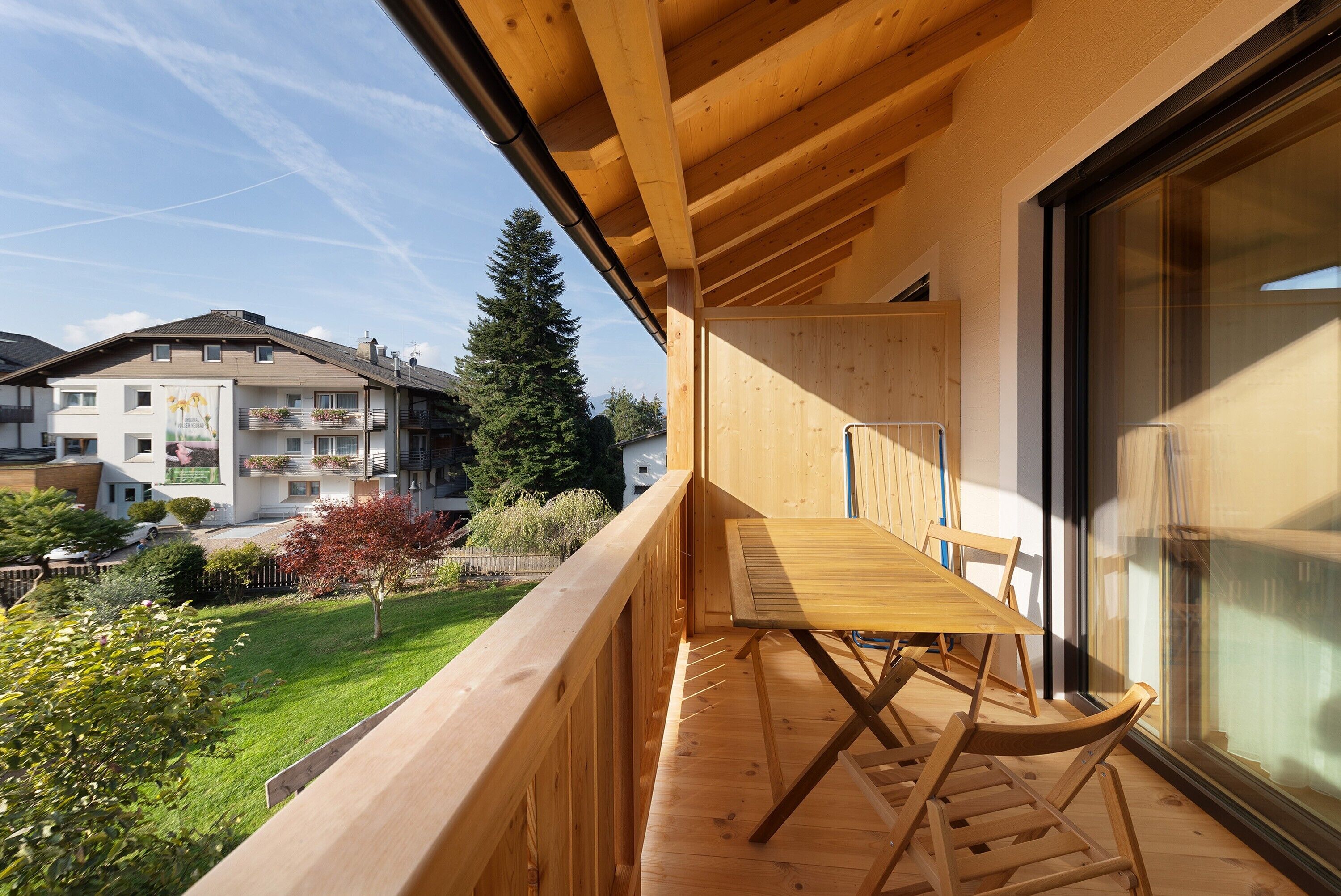 Ein Holzbalkon mit Tisch und Stühlen, mit Blick auf einen Garten und angrenzende Gebäude.