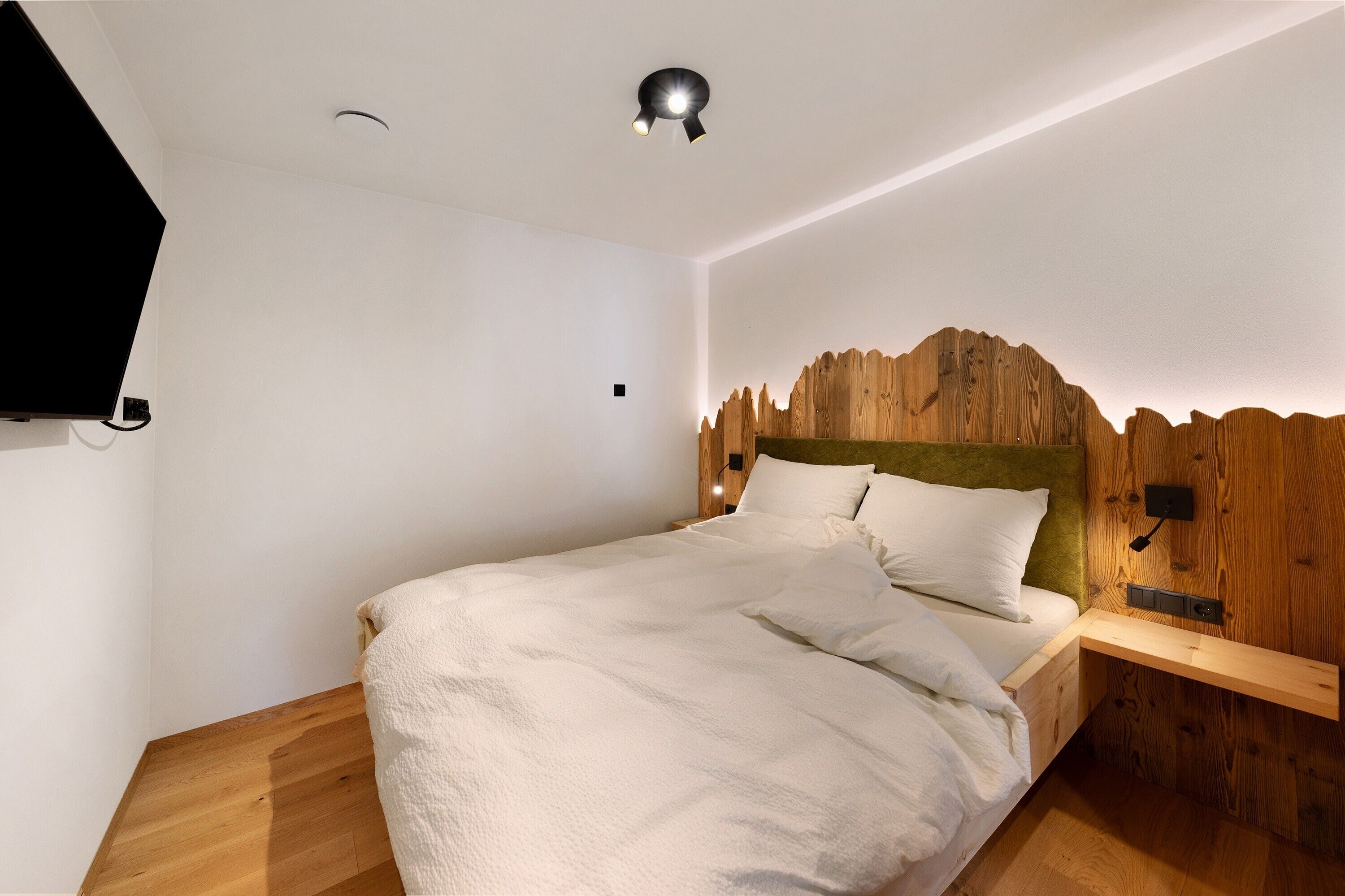 Une chambre à coucher avec un lit, un téléviseur fixé au mur, une tête de lit en bois et une table de chevet.