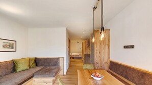 Un intérieur moderne avec un banc en bois, une table à manger sur laquelle se trouve un saladier rempli de fruits, et un couloir menant à une chambre.