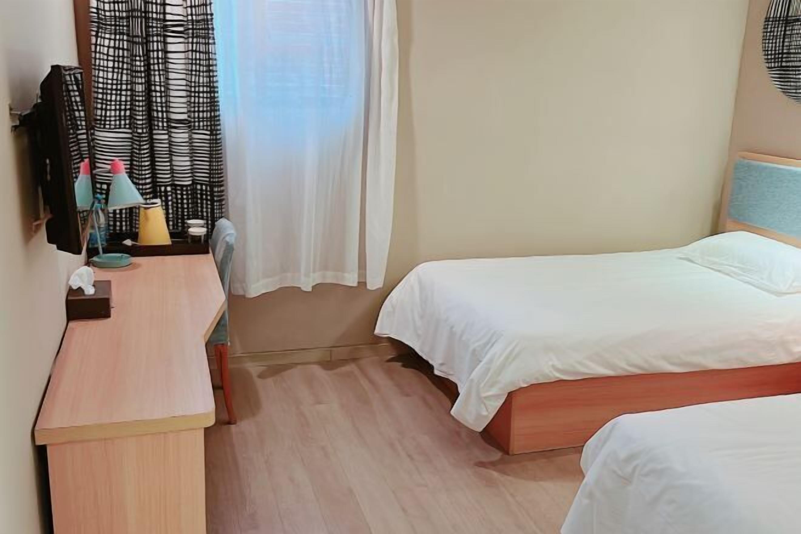 A hotel room with two beds, a desk, a chair, a small table with a lamp, and a window with curtains.