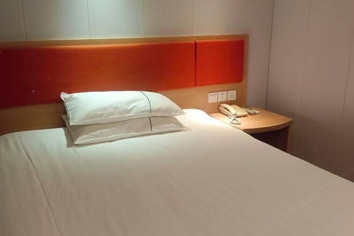 A hotel room with a bed, pillows, a bedside table with a telephone, and a wall socket.