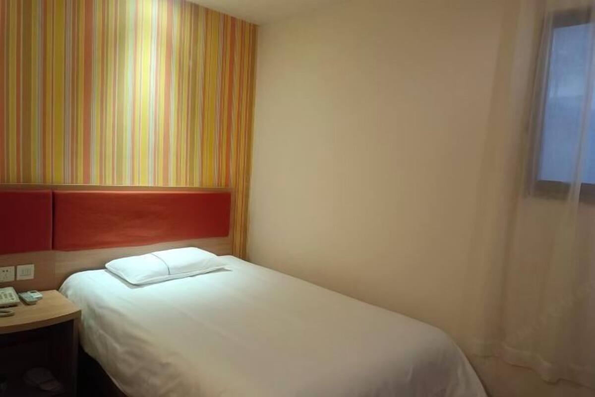 A single bed with a white mattress and pillow, a wooden nightstand with a telephone, and a striped wall behind the bed.