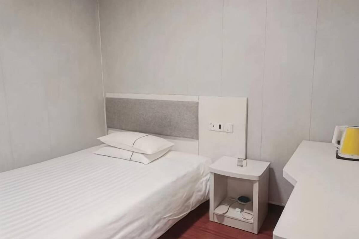 A single bed with a white mattress and pillow, a bedside table with a lamp, and a small shelf with a cup and a book.