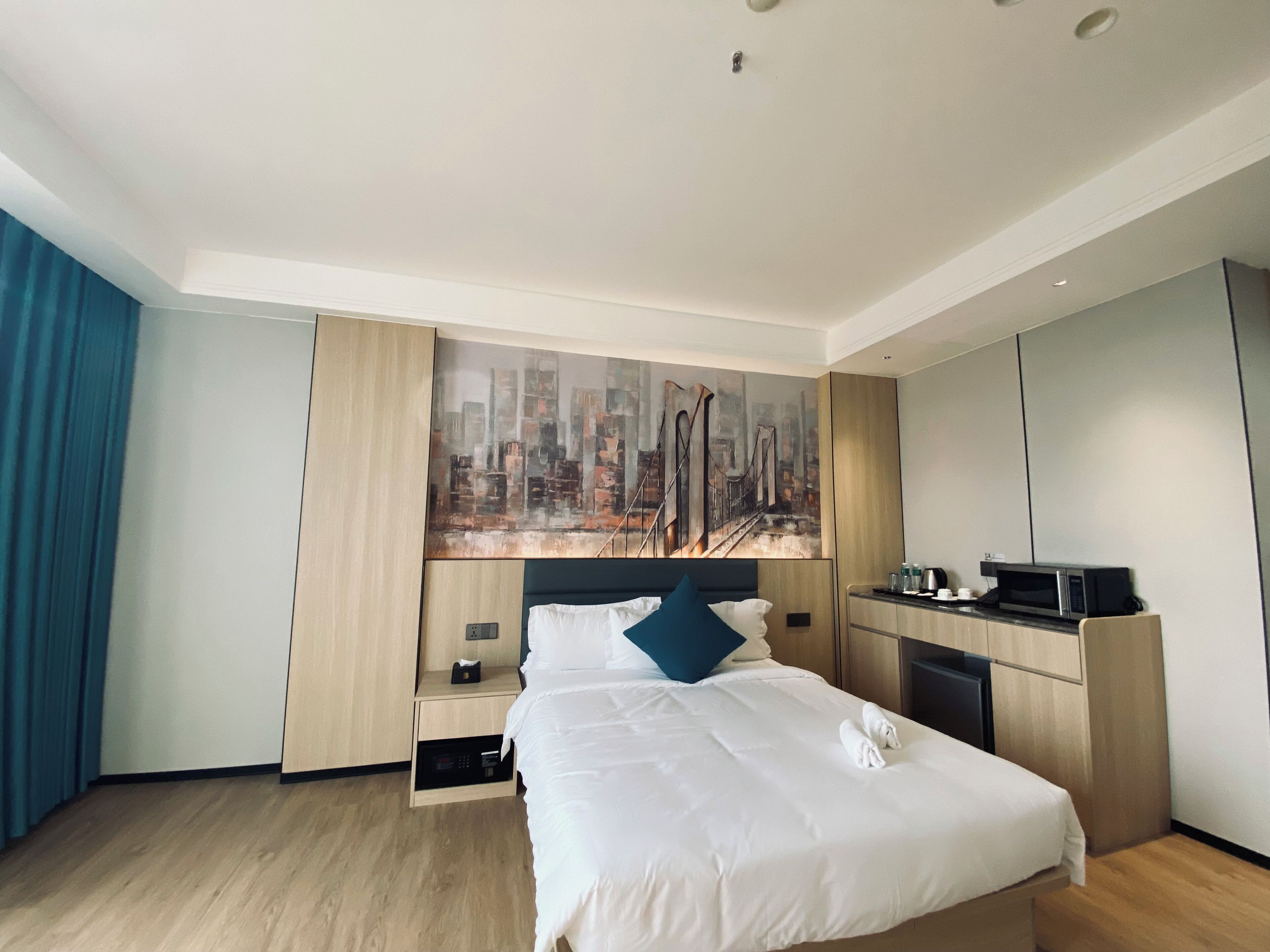 Habitación de hotel con cama, escritorio con microondas, un cuadro de paisaje urbano en la pared y una cortina azul.