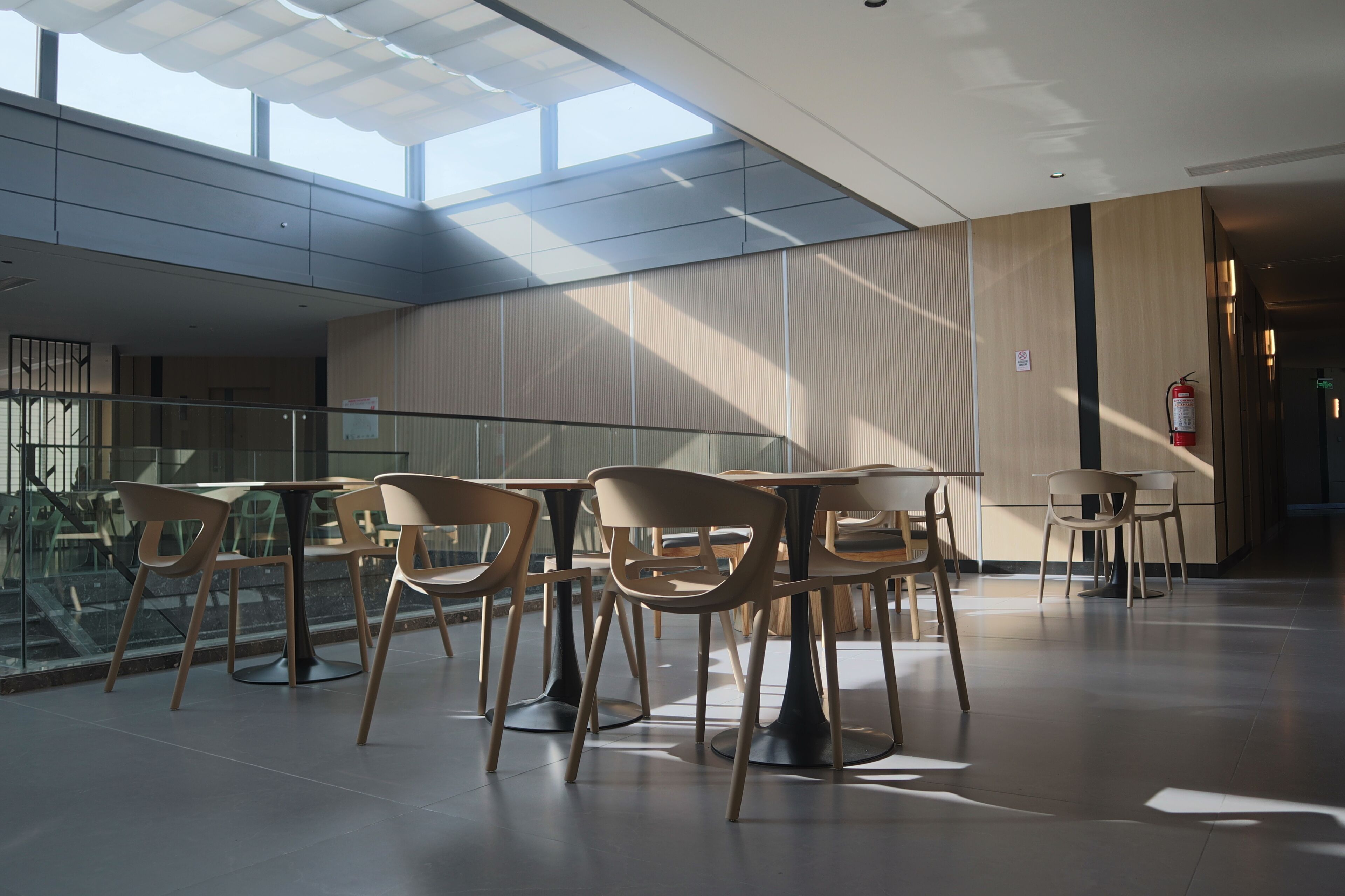 A modern indoor space with natural lighting, featuring a seating area with tables and chairs, a fire extinguisher, and a visible staircase.
