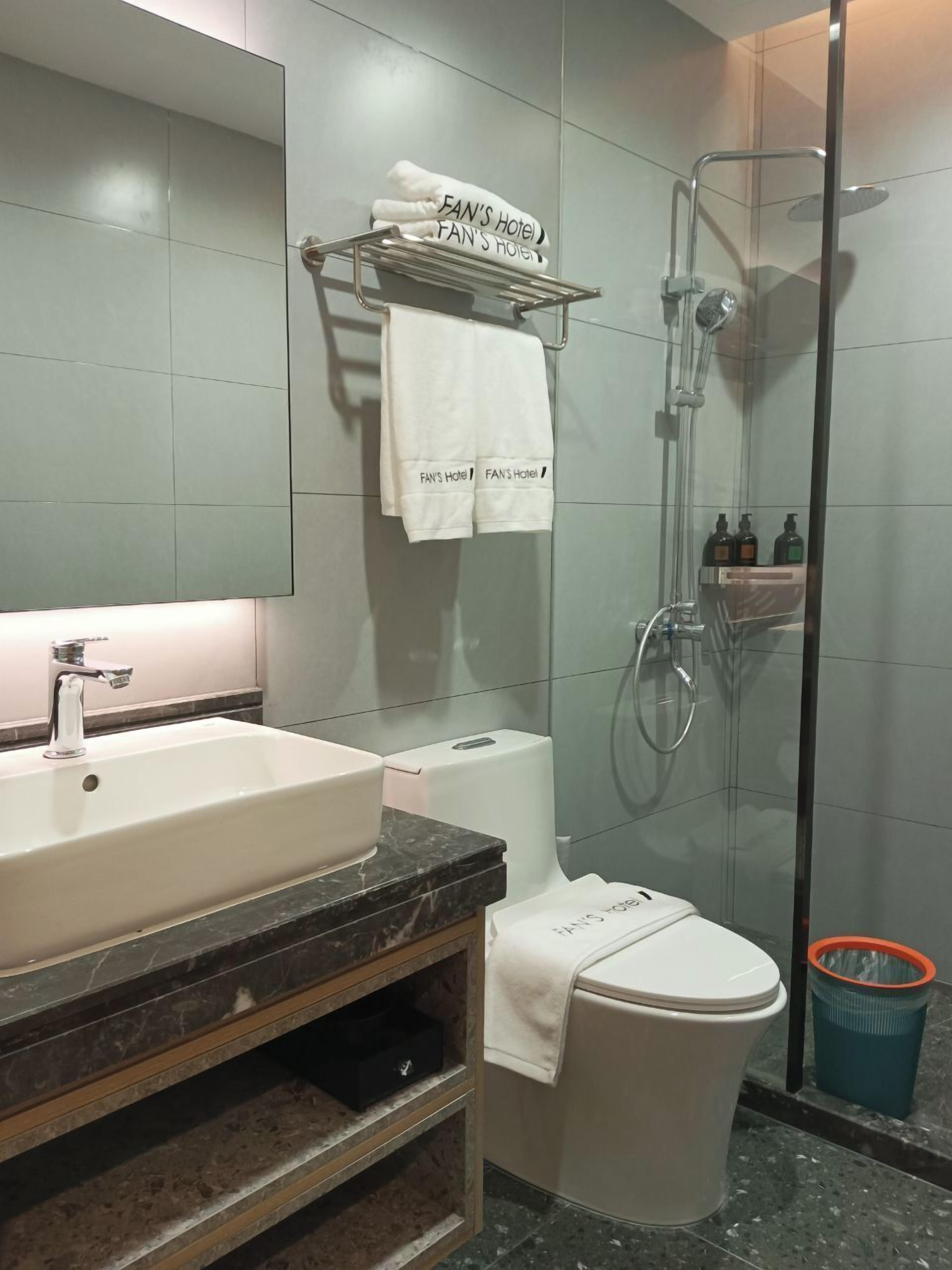 Une salle de bain moderne avec un grand lavabo, des toilettes, une douche en verre et un porte-serviettes sur lequel sont disposées des serviettes étiquetées « Hôtel FAN’S ».