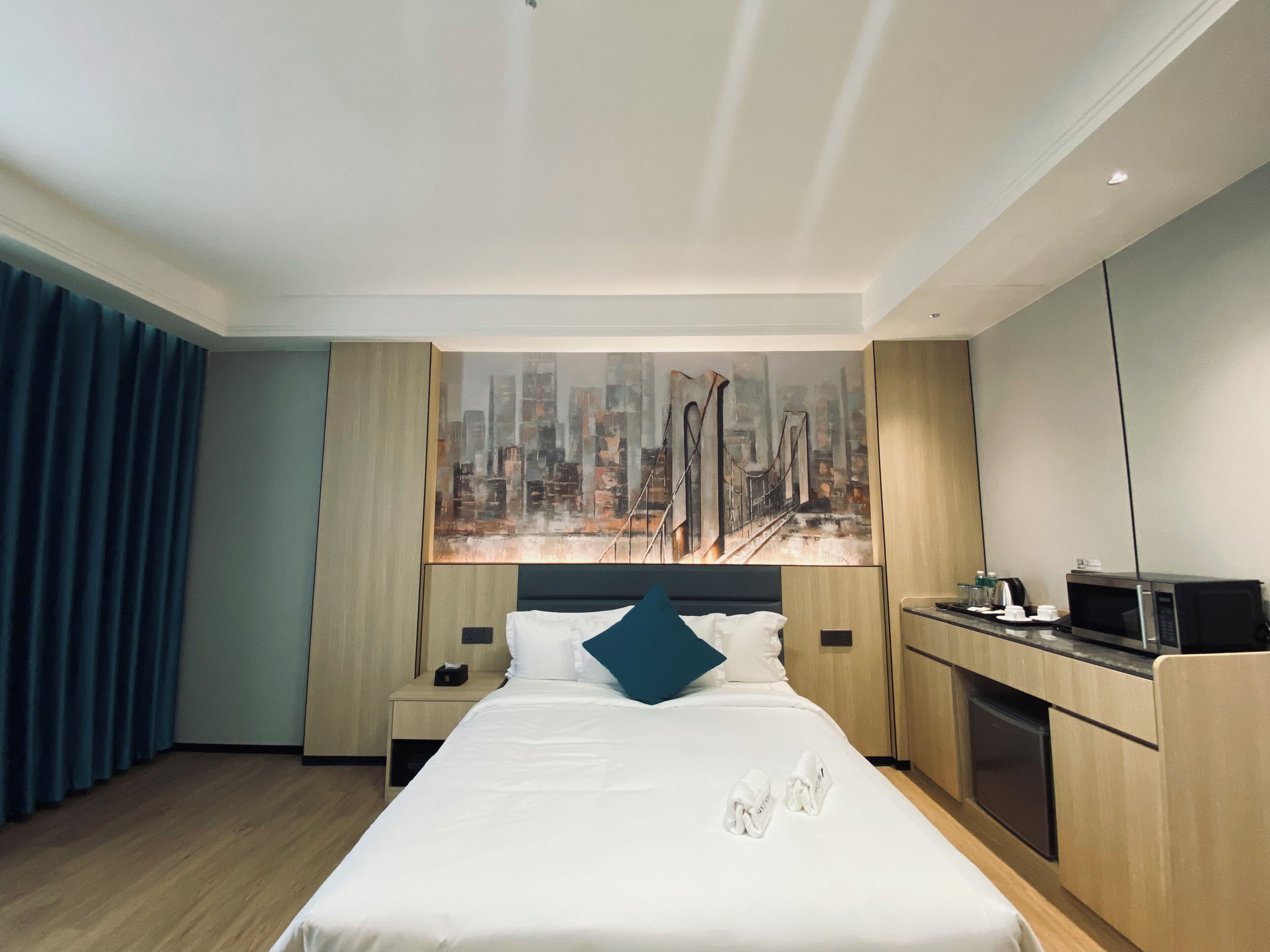 Habitació d'hotel amb un llit, una taula de treball, un microones i un quadre de paisatge urbà a la paret.