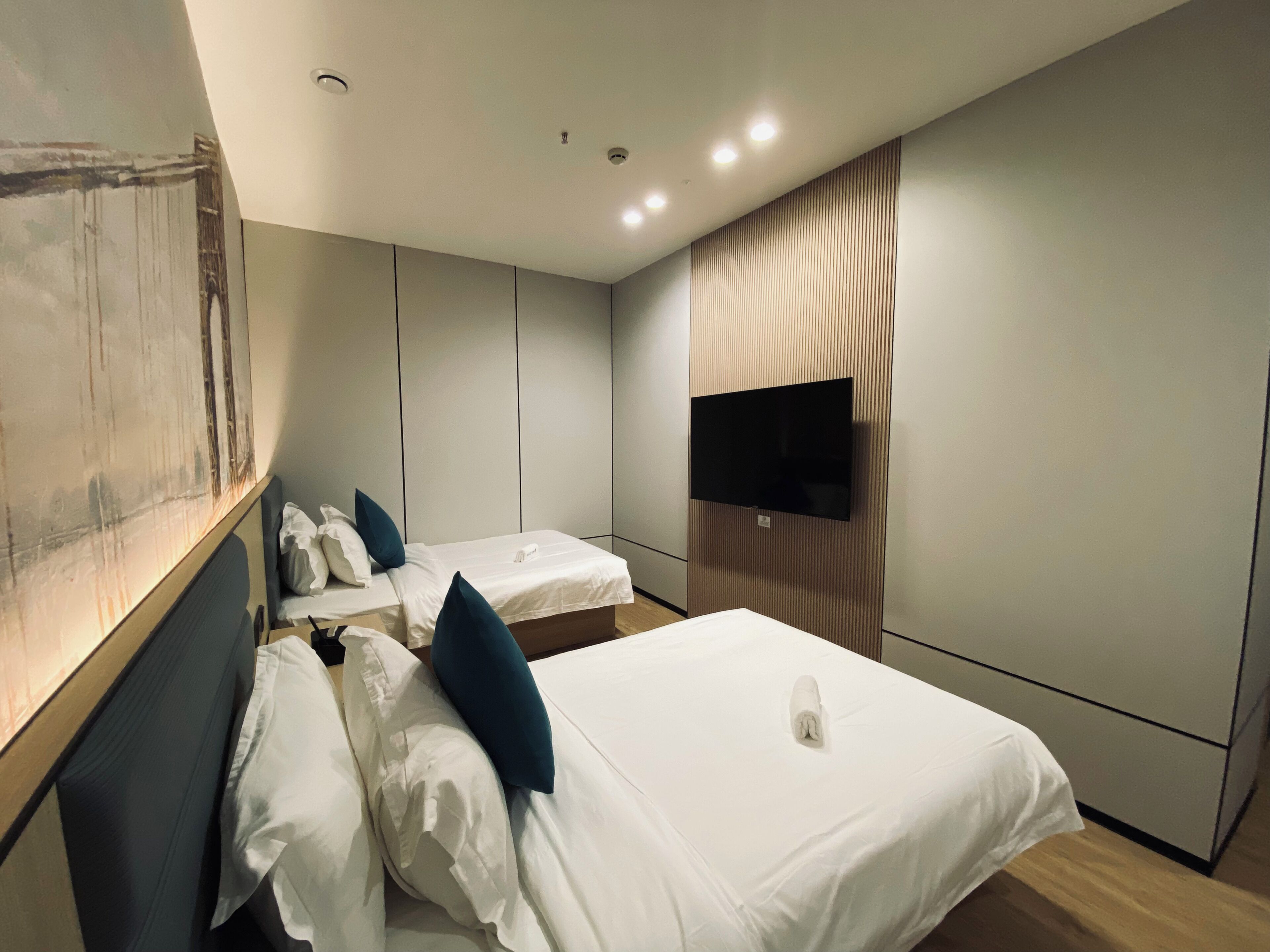 A hotel room with two beds, a flat-screen TV mounted on the wall, and a painting on the left wall.