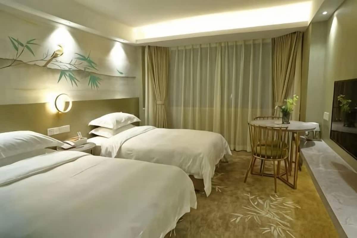 A hotel room with two beds, a round table with chairs, a wall-mounted lamp, and a large window with curtains.