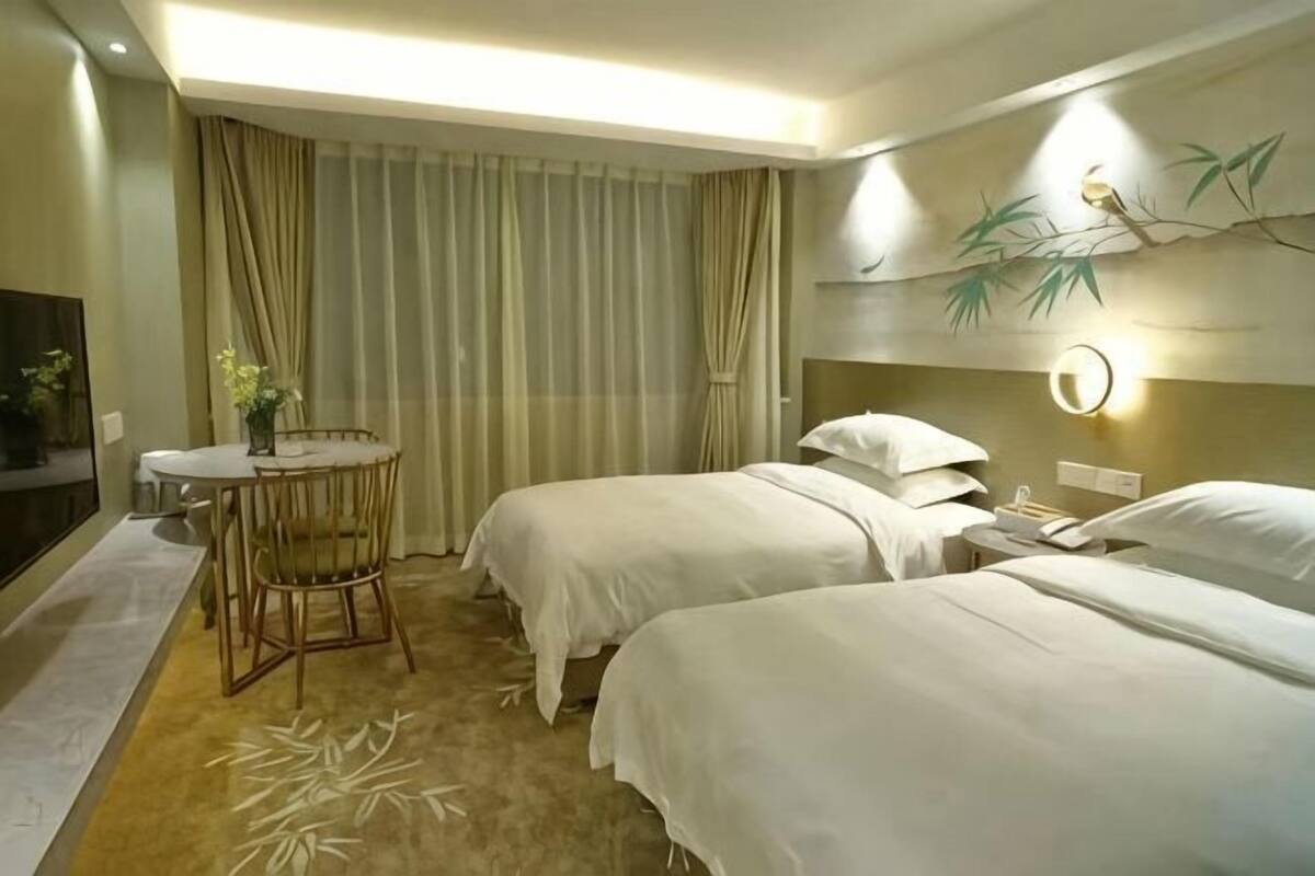 A hotel room with two beds, a small table with a chair, a round mirror, and a wall-mounted lamp.