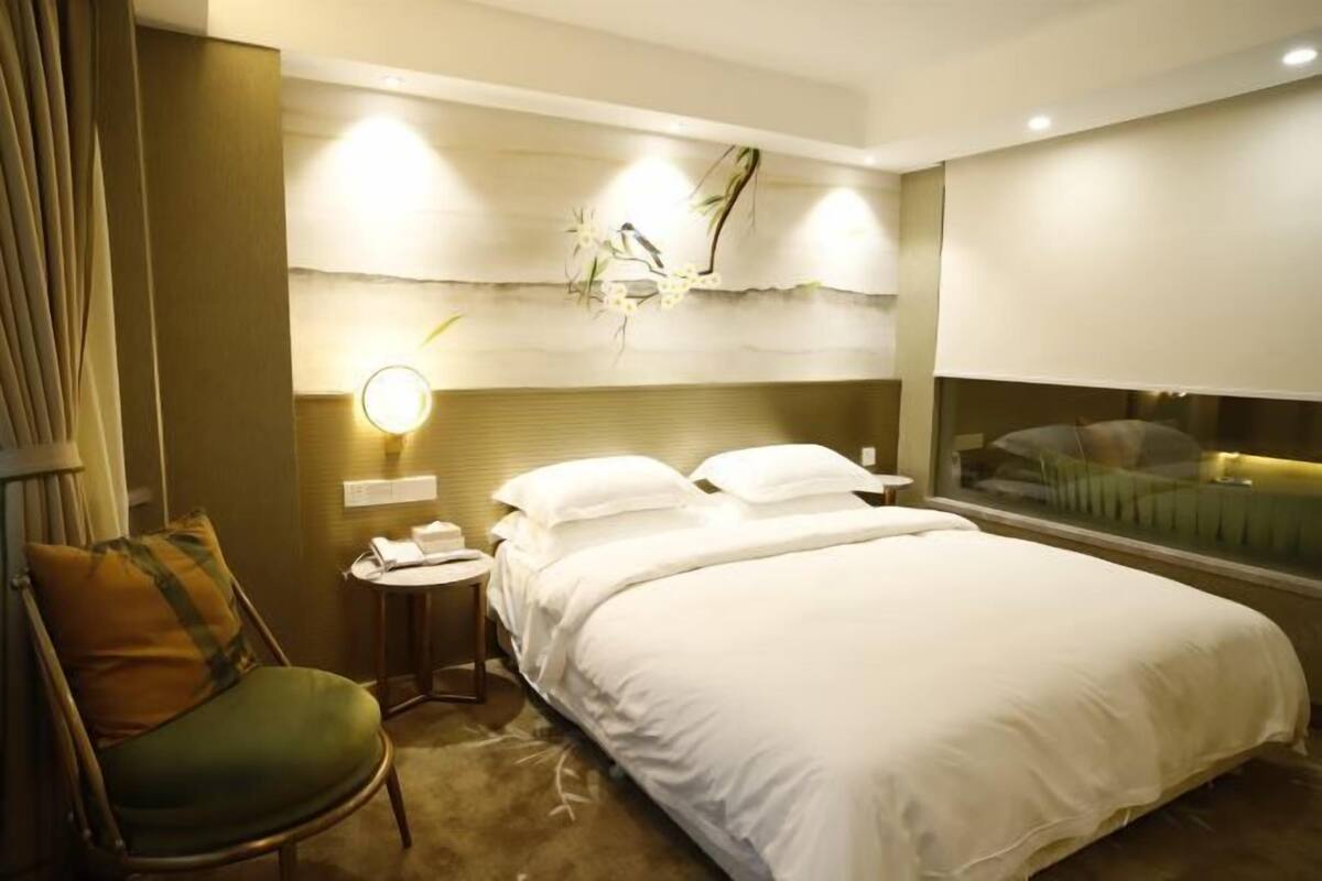 A hotel room with a bed, a chair, a bedside table, a wall-mounted lamp, and a window.