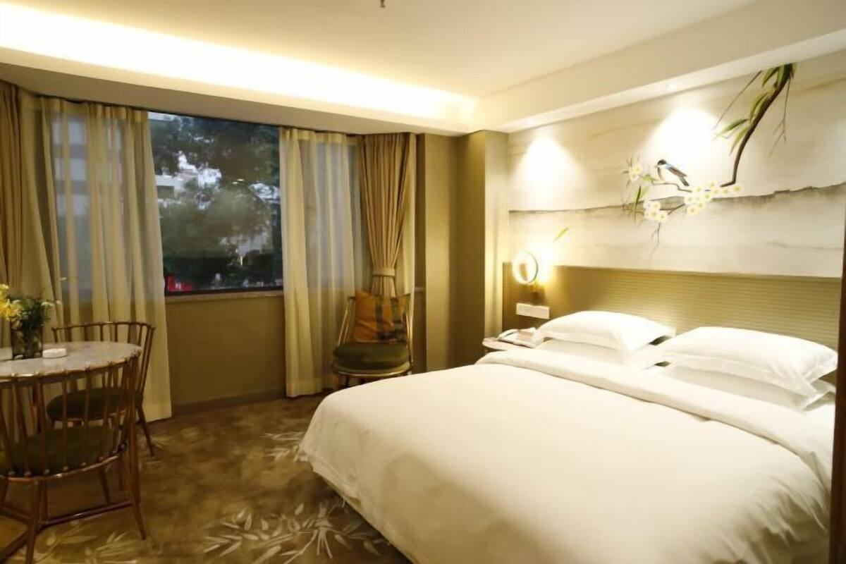 A hotel room with a large bed, a dining table with chairs, a chair, a lamp, and a window with curtains.