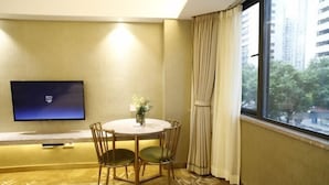 A hotel room with a round table, two chairs, a flat-screen TV mounted on the wall, a window with curtains, and a view of buildings and trees outside. - Cui Zhu Hotel (Shenzhen)