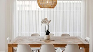 Un coin repas avec une table en bois, des chaises blanches, un luminaire suspendu en fibres tressées et une plante orchidée en pot.