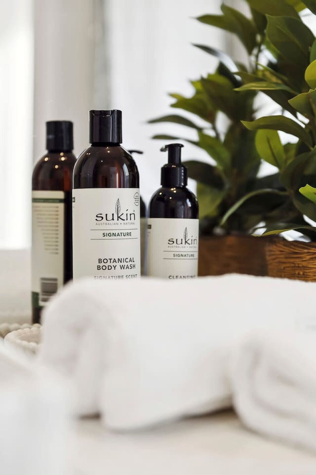 A bathroom with a white towel, a bottle of Sukin Signature Botanical Body Wash, and a bottle of Sukin Signature Cleanse.