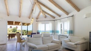 A spacious living room with a high ceiling and wooden beams, a large white sofa, a dining area with chairs, and a view of the outdoors. - Villa 'Amata' with Sea View, Wi-Fi and Air Conditioning (Arzachena)