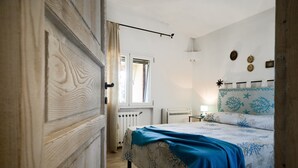 A bedroom with a wooden door, a bed with a blue bedspread, a radiator, and a window with curtains. - Villa 'Amata' with Sea View, Wi-Fi and Air Conditioning (Arzachena)