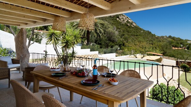 An outdoor dining area with a wooden table and wicker chairs, overlooking a pool and greenery. - Villa 'Amata' with Sea View, Wi-Fi and Air Conditioning (Arzachena)