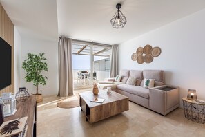 Un salon moderne avec un canapé, une table basse et un téléviseur. On y trouve également une plante, une lampe et une vue sur une terrasse extérieure.