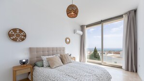Un dormitorio con una cama grande, una mesita de noche con una lámpara, aire acondicionado instalado en la pared y vistas a la ciudad a través de un ventanal amplio.