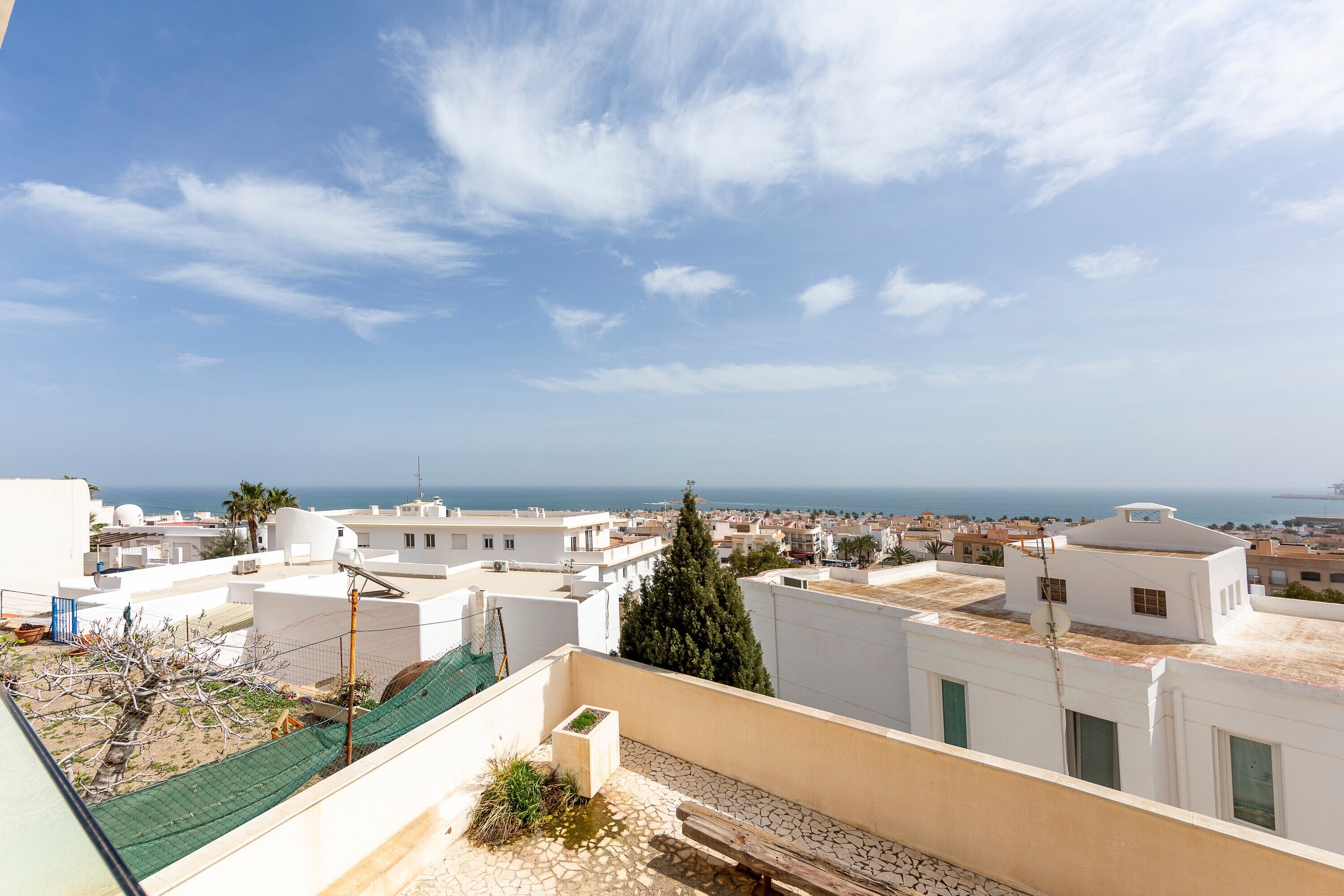 A rooftop terrace with a view of the sea, white buildings, and a palm tree.