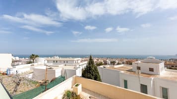 A rooftop terrace with a view of the sea, white buildings, and a palm tree.