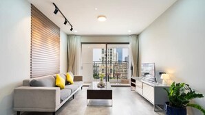 A modern living room with a sofa, coffee table, and a TV on a stand. There is a balcony with a view of buildings. - Saigon Royal - Trendy Modern Apartment (Ho Chi Minh City)