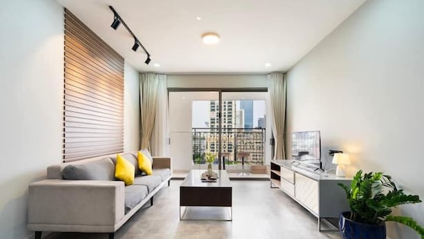 A modern living room with a sofa, coffee table, and a TV on a stand. There is a balcony with a view of buildings.