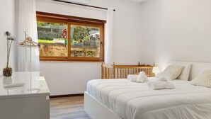 A bedroom with a bed, a dresser, a window with a view of greenery, and a white wall.