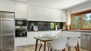 A modern kitchen with a built-in oven, microwave, and dishwasher, a dining table with chairs, and a window with a view of trees.