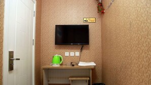 Habitación de hotel con televisión montada en la pared, un pequeño escritorio con hervidor, un cubo de basura y una puerta con manija.